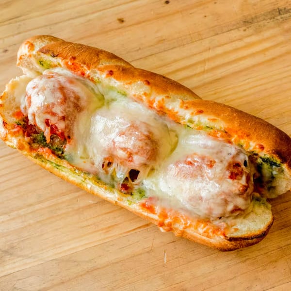 Totally Baked Pizza's meatball sub, topped with melted cheese and pesto, on a wooden surface.