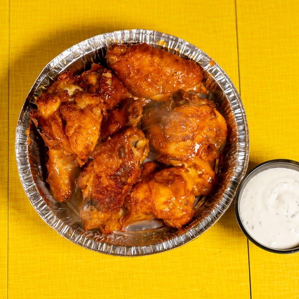 Steaming hot chicken wings in a foil container with a side of ranch dressing on a yellow surface.