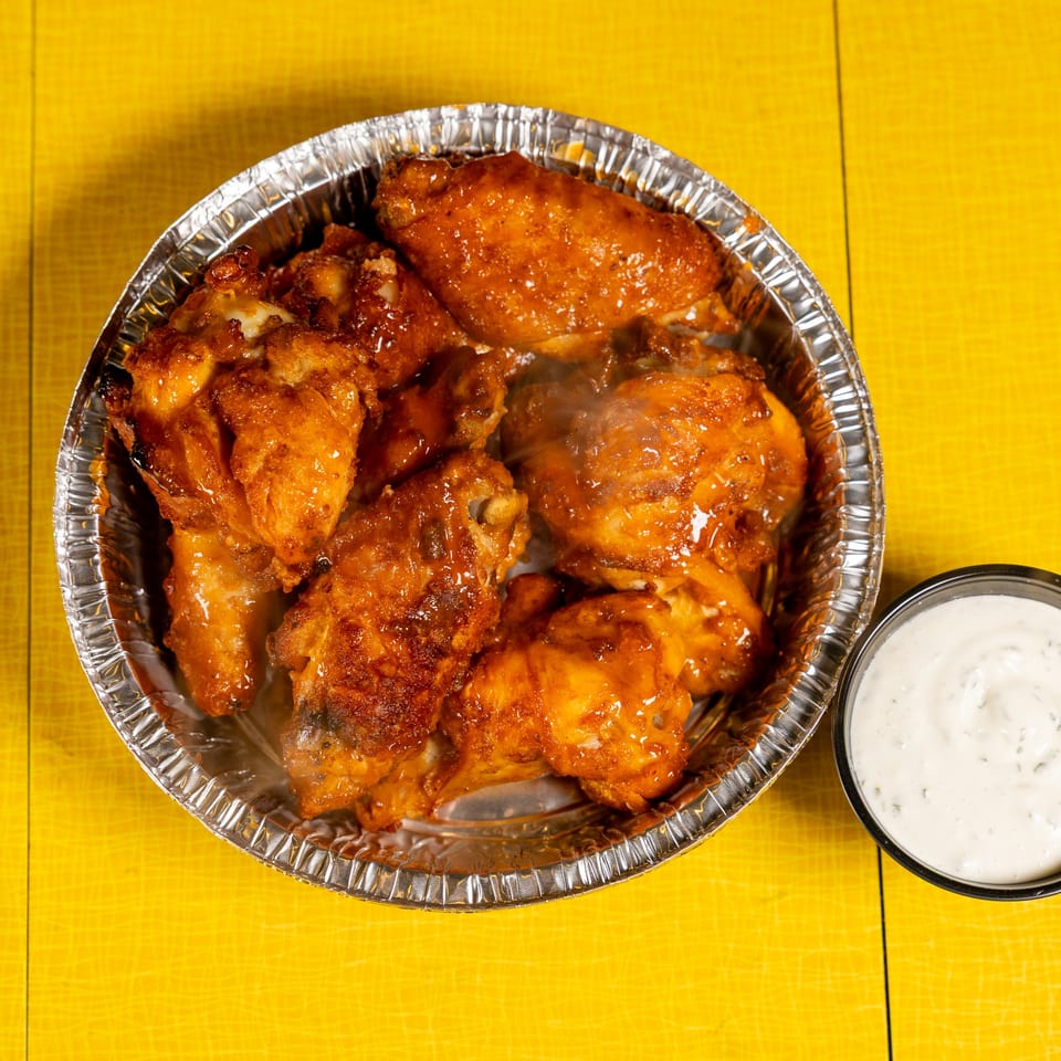 Steaming hot chicken wings in a foil container with a side of ranch dressing on a yellow surface.