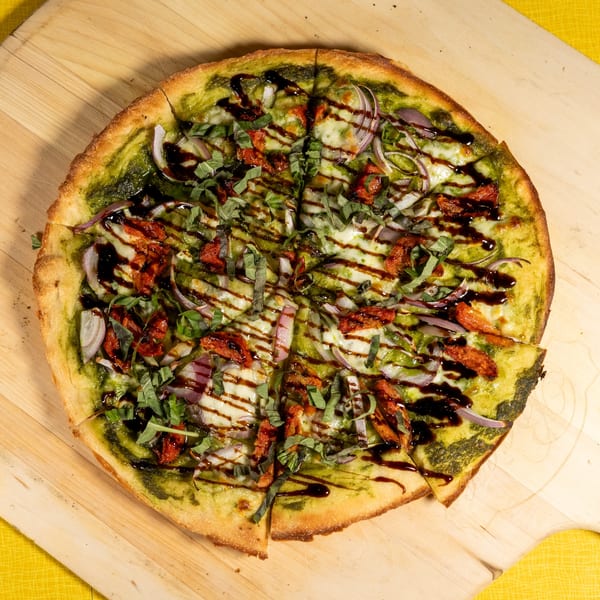 Pesto pizza with sun-dried tomatoes, red onion, mozzarella, basil, and balsamic glaze on a wooden peel.