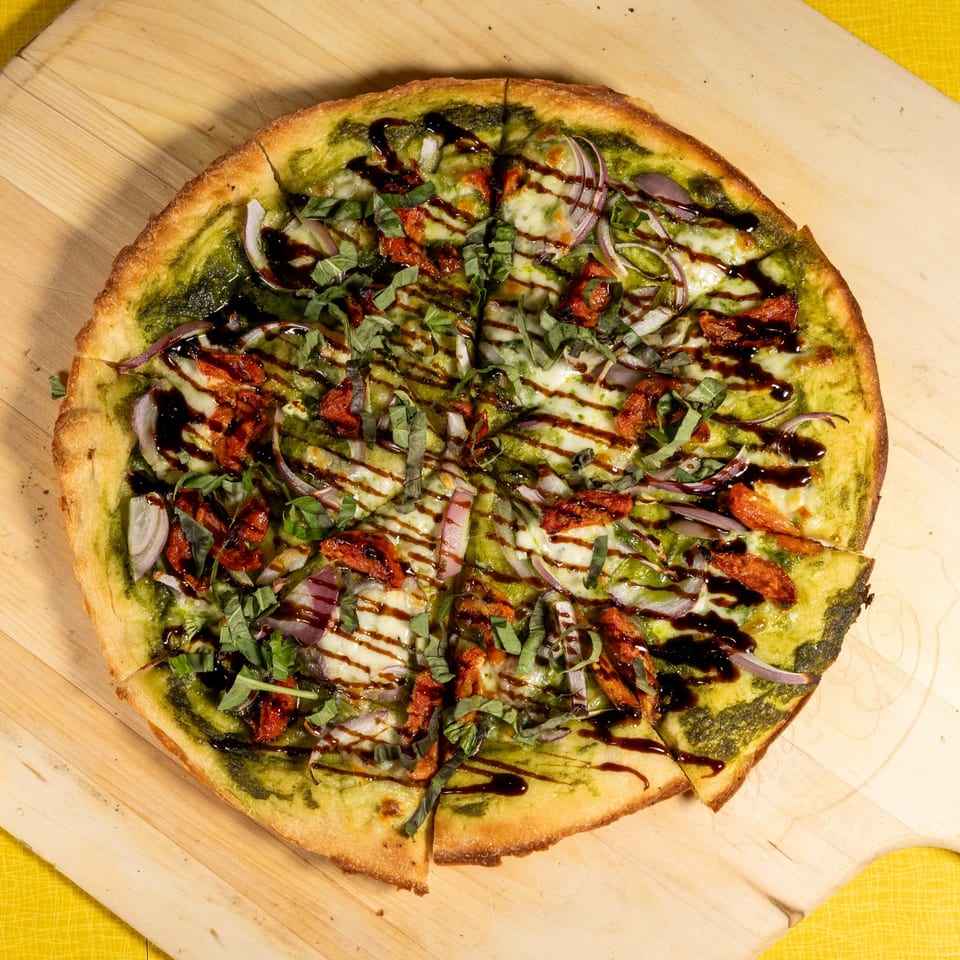 Pesto pizza with sun-dried tomatoes, red onion, mozzarella, basil, and balsamic glaze on a wooden peel.