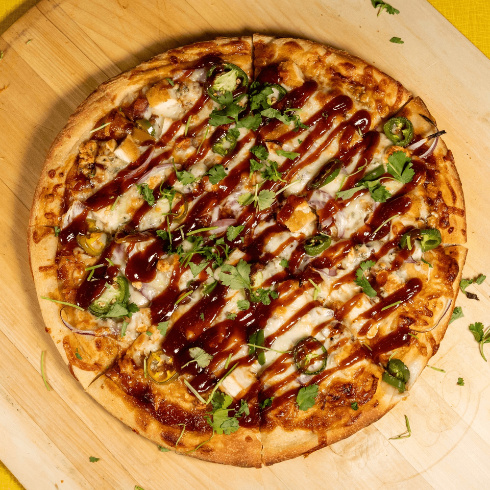 BBQ chicken pizza with cilantro, red onions, jalapeños, and a BBQ sauce drizzle on a wooden board.
