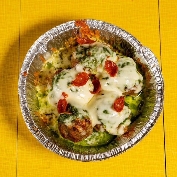 Meatballs covered in melted cheese, green pesto, and pepperoni slices in a foil container.