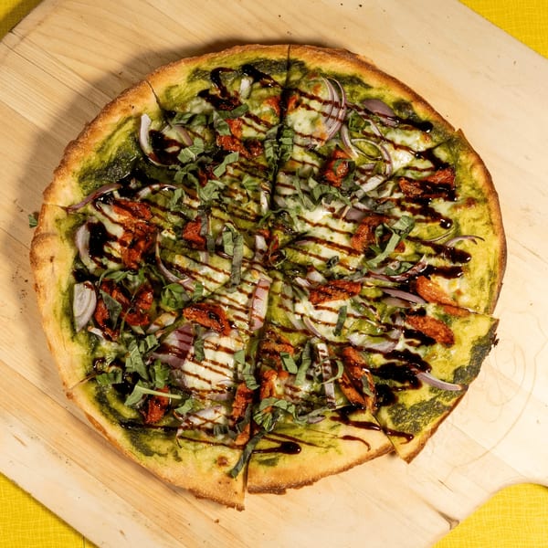 Pesto pizza with sun-dried tomatoes, red onion, mozzarella, and balsamic glaze on a wooden peel.