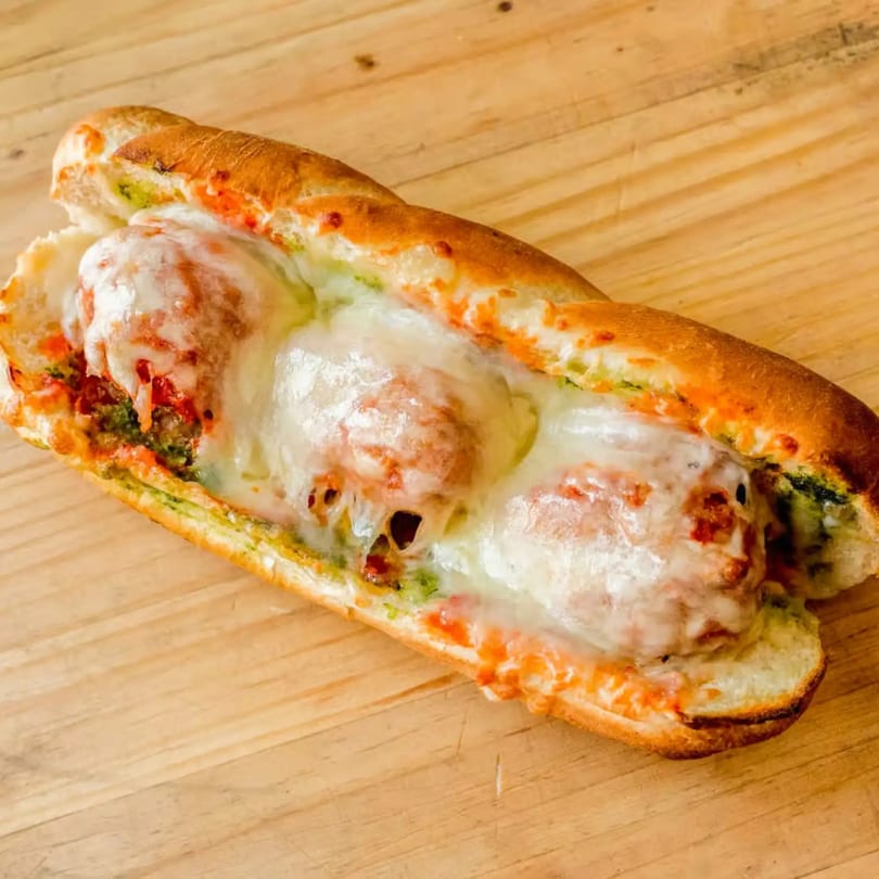 A Totally Baked Pizza meatball sub with melted cheese, red sauce, and green pesto on a wooden surface.