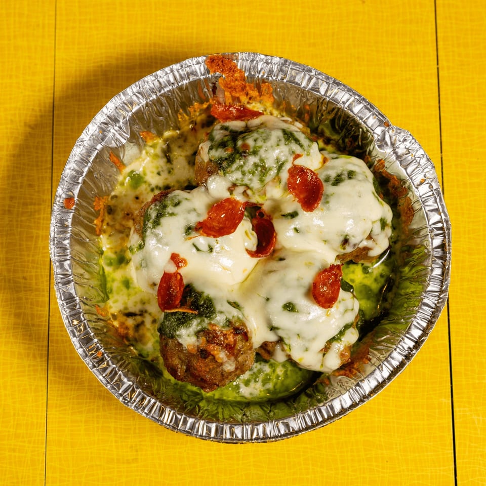 Meatballs covered in melted cheese, green sauce, and pepperoni slices in a foil container.