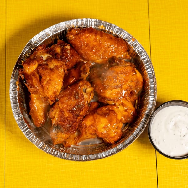 Steaming hot chicken wings in a foil container with a side of ranch dressing on a yellow table.