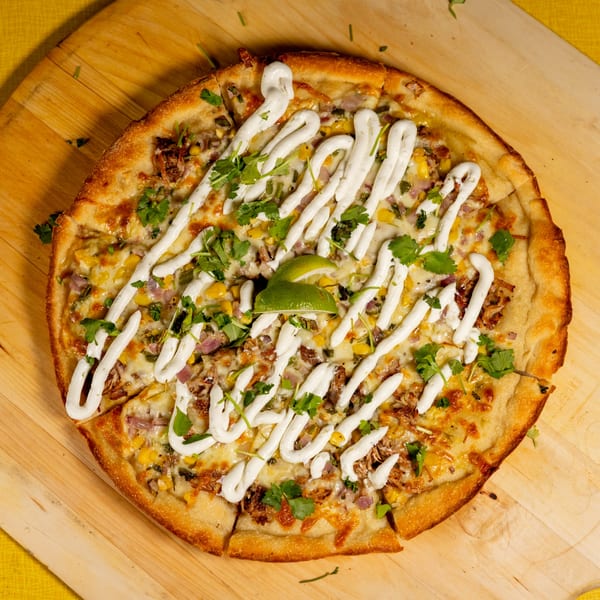 A whole pizza with white sauce, corn, cilantro, and lime wedges on a wooden board.