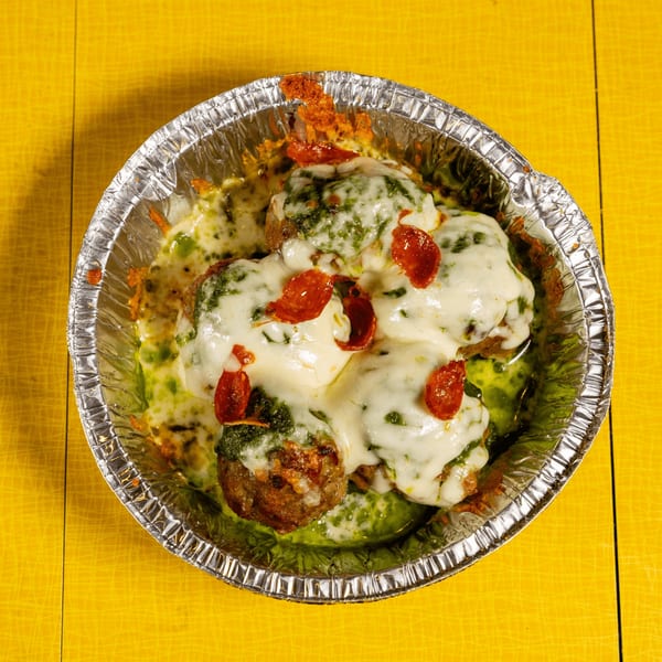 Meatballs covered in melted cheese, green pesto, and pepperoni slices in a foil container.