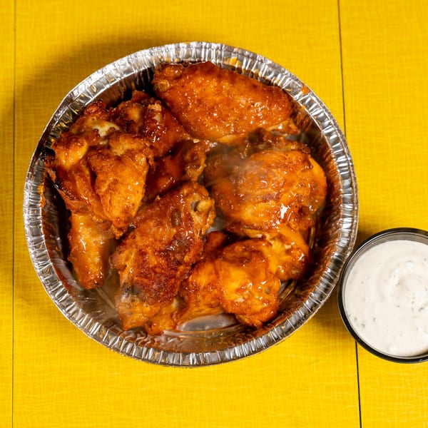 Steaming hot chicken wings in a foil container with a side of ranch dressing on a yellow table.